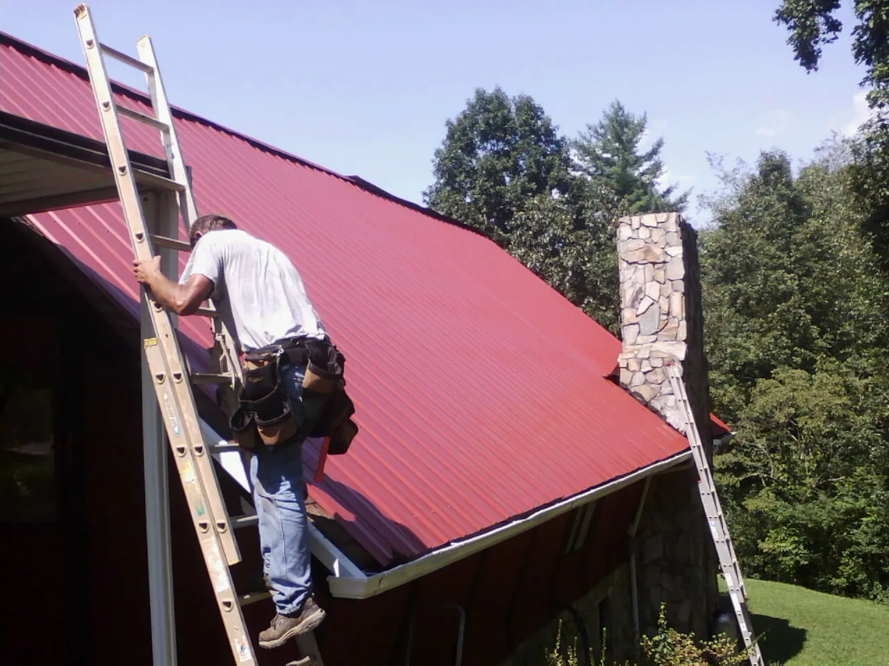 Roofer on a ladder beside a red metal roof and stone chimney — Soffit & Fascia Repair by Roofing Brothers in Bernardsville